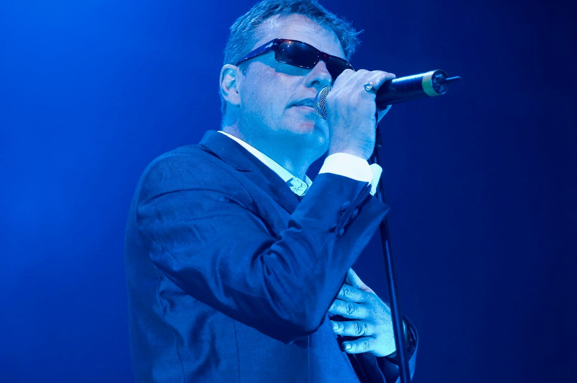 A Madness band member sings into a microphone in close-up under blue stage light, wearing dark sunglasses and a black suit jacket over a white shirt.