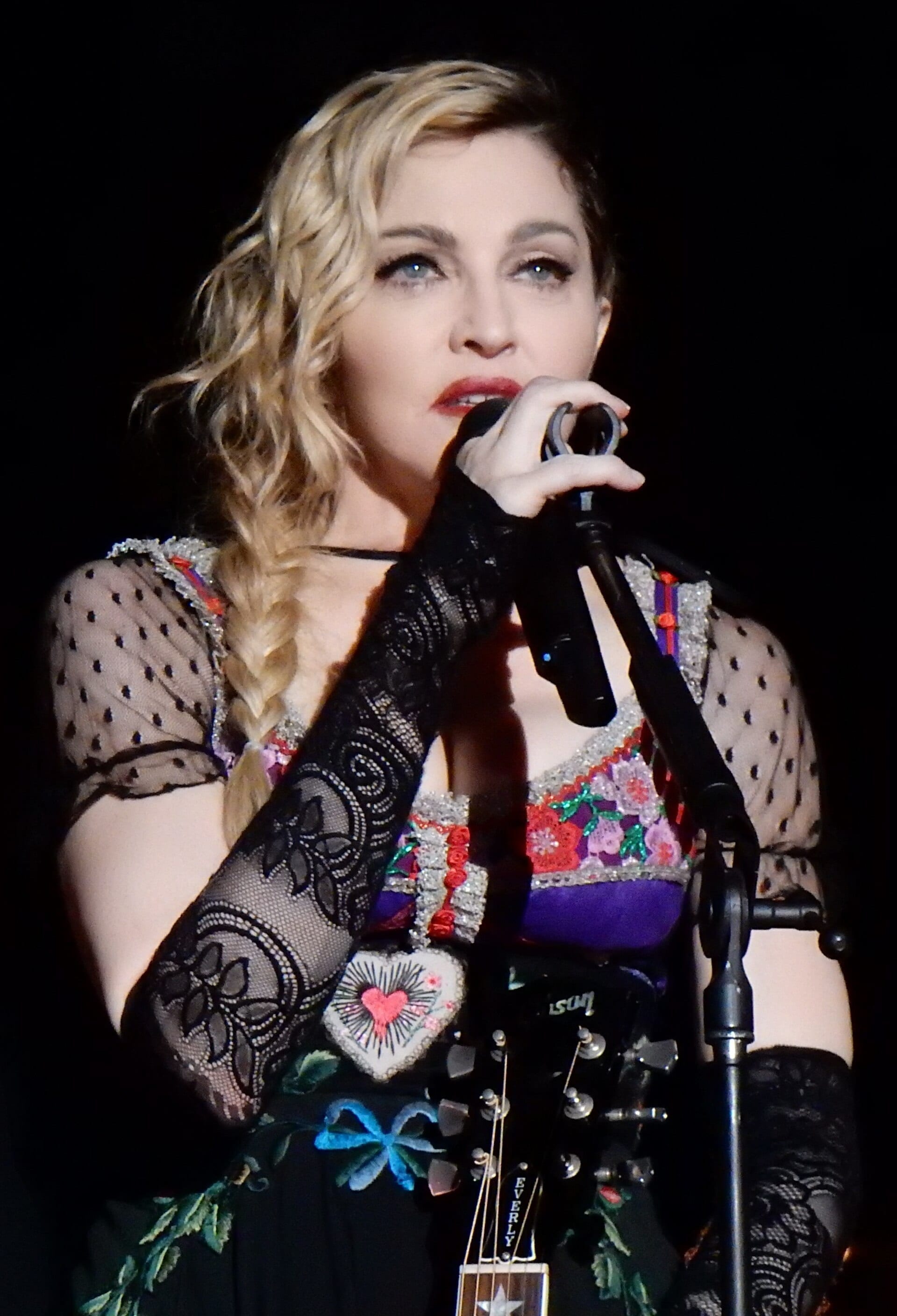 Madonna sings into a handheld microphone in a lace and floral-embroidered stage costume with sheer polka-dot sleeves, blonde hair in a side braid, holding the neck of a ukulele.