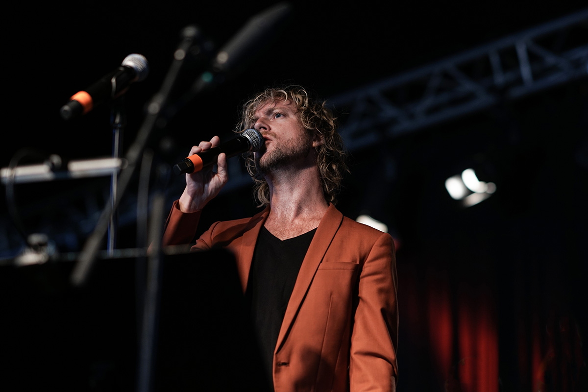 Mads Mathias sings into a handheld microphone in a rust-orange blazer over a black top, mic stands and lighting truss behind him.