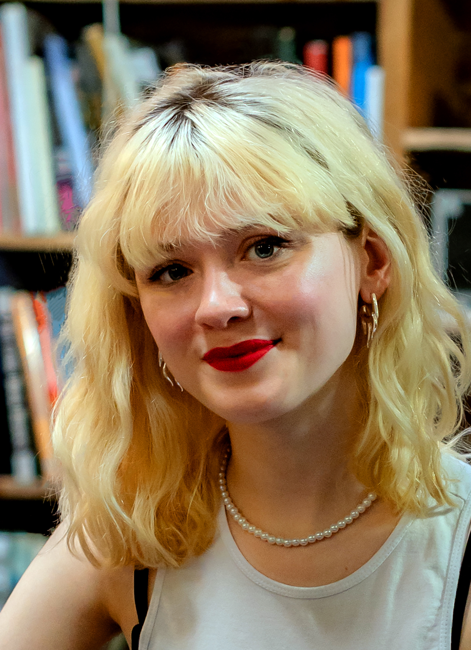 Maisie Peters smiles at the camera in a white vest top and a pearl necklace, blonde hair with a fringe, in front of packed record-shop shelves.
