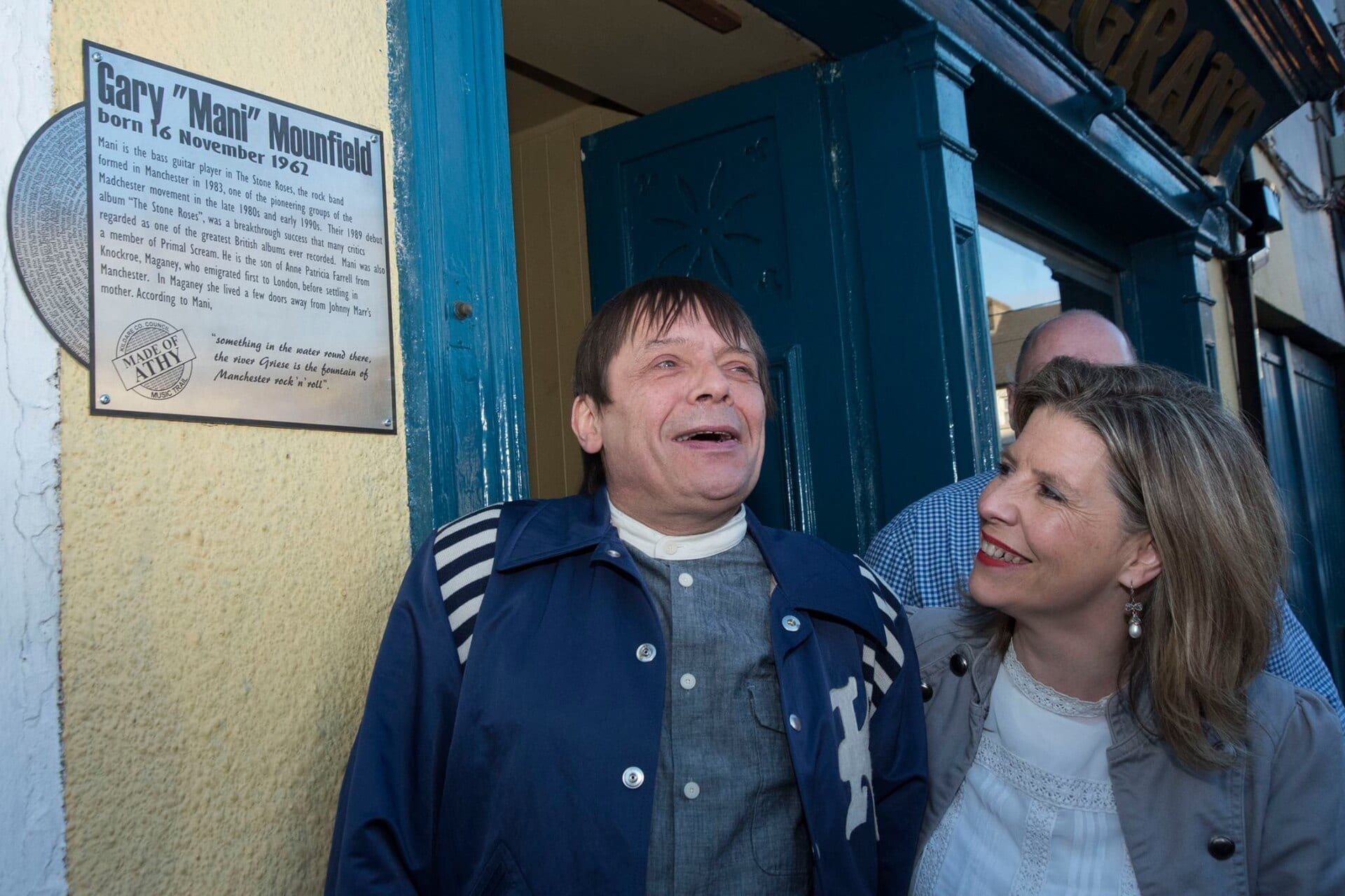 Mani, wearing a striped-collared navy jacket over a grey shirt, smiles and looks up while standing next to a woman in a doorway, beside a plaque reading "Gary 'Mani' Mounfield, born 16 November 1962" that credits him as the Stone Roses and Primal Scream bassist.