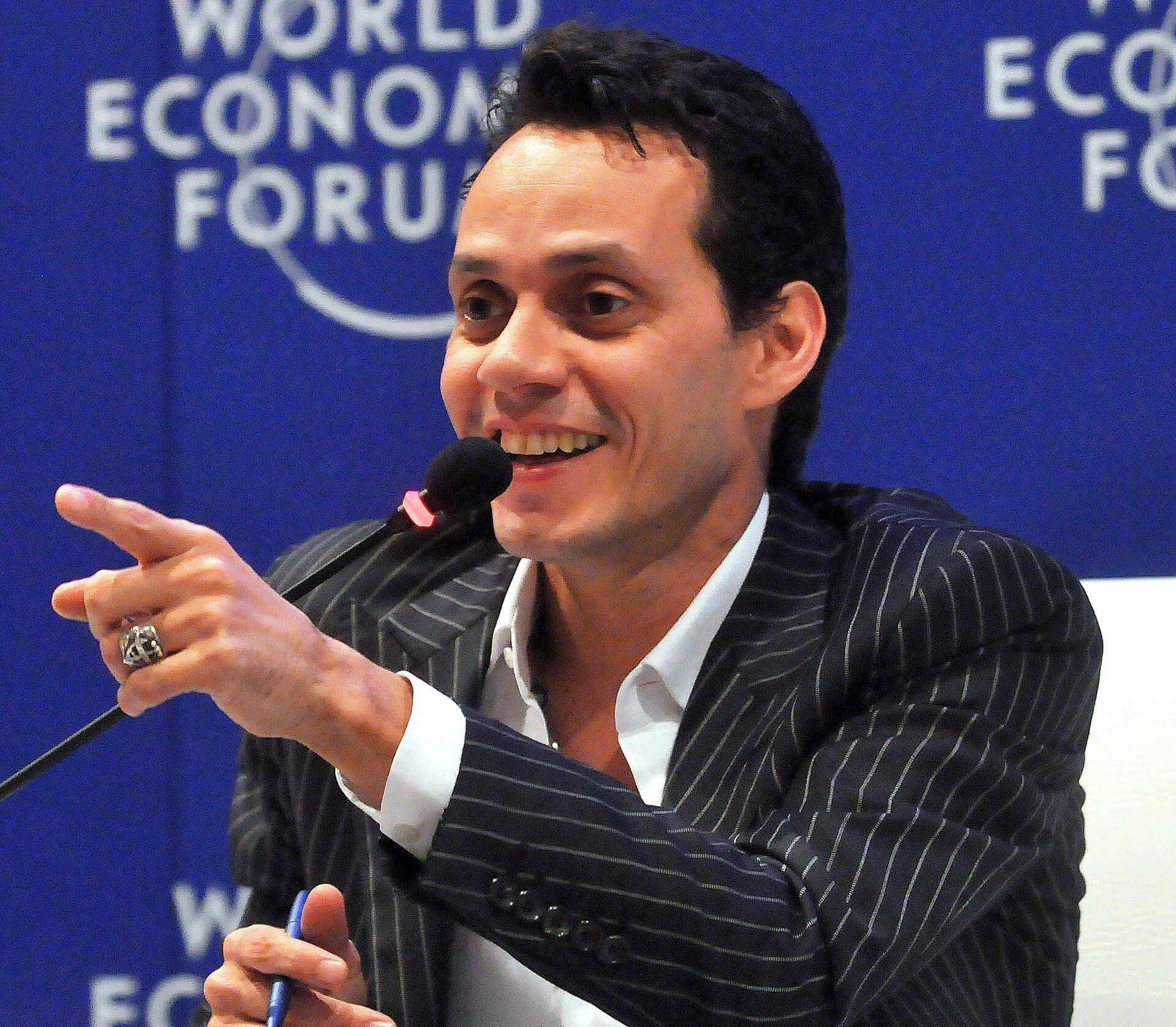 Marc Anthony smiles and points while speaking into a microphone, wearing a pinstriped suit in front of a World Economic Forum backdrop.