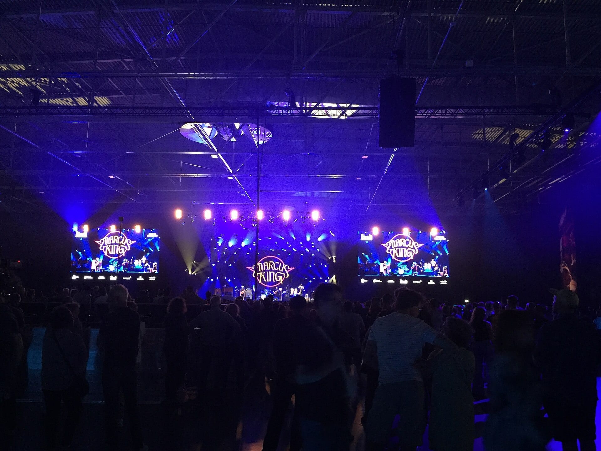 A wide, purple-lit concert hall shows the Marcus King Band performing on a distant stage below video screens displaying his round logo.