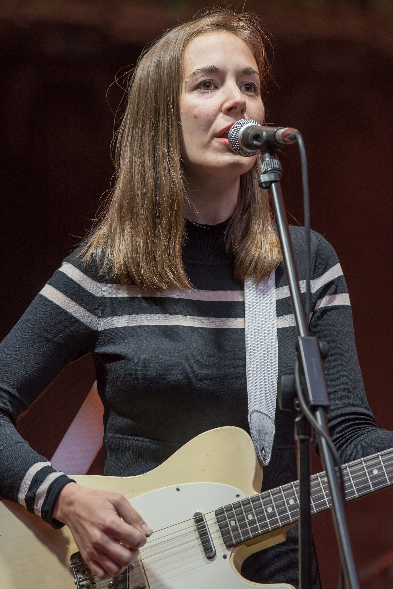 Margaret Glaspy sings into a microphone while playing an electric guitar, wearing a striped black and gray sweater.