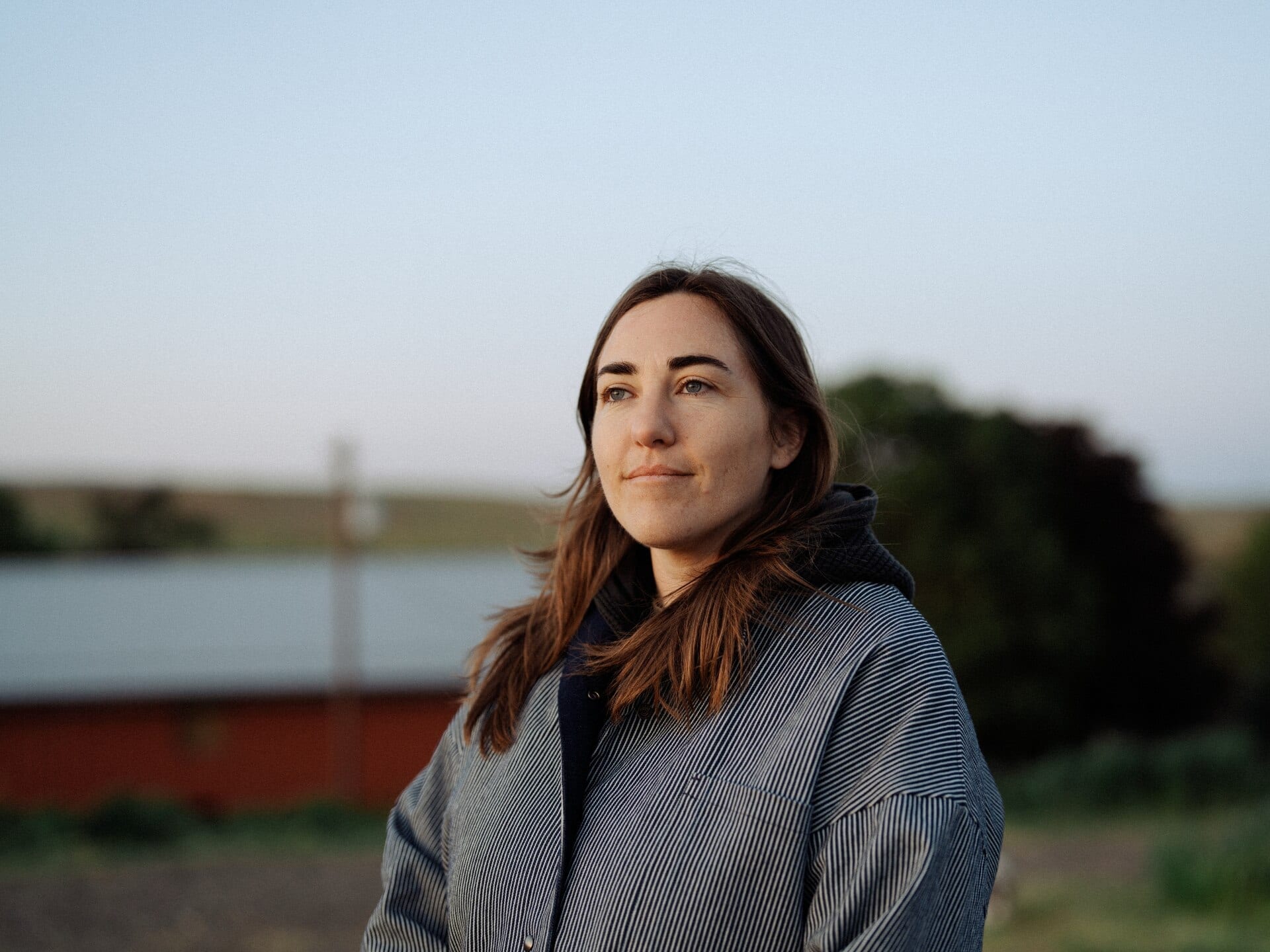 Margo Cilker stands outdoors at dusk in a striped jacket, looking off to the side with open farmland and a barn behind her.