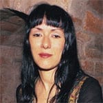 A close portrait of Maria Gabriela Epumer, a woman with dark hair and blunt bangs, standing against a stone wall.