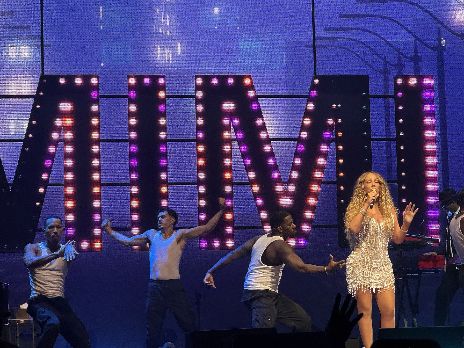 Mariah Carey sings at the edge of a lit arena stage in a sparkling outfit, dancers behind her and large illuminated letters at the back of the set.