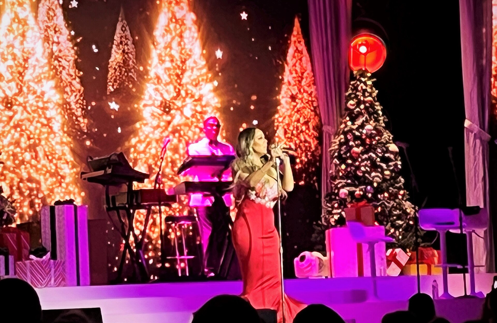 Mariah Carey in a long red dress singing into a mic stand on a stage dressed with Christmas trees, wrapped presents and a glittering backdrop, a keyboard player behind her