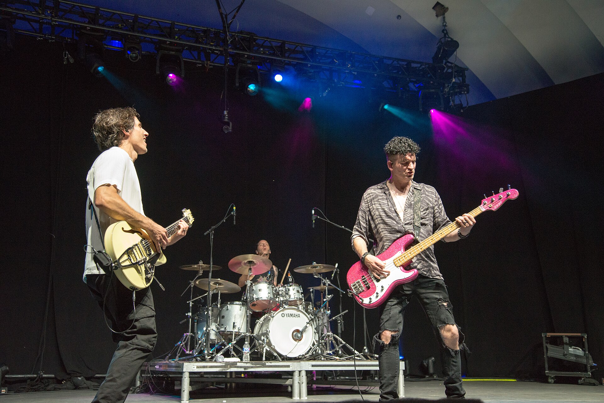 Marianas Trench on a large stage, a guitarist with a cream hollow-body on the left, a bassist with a pink sparkle bass on the right and the drummer on a riser between them