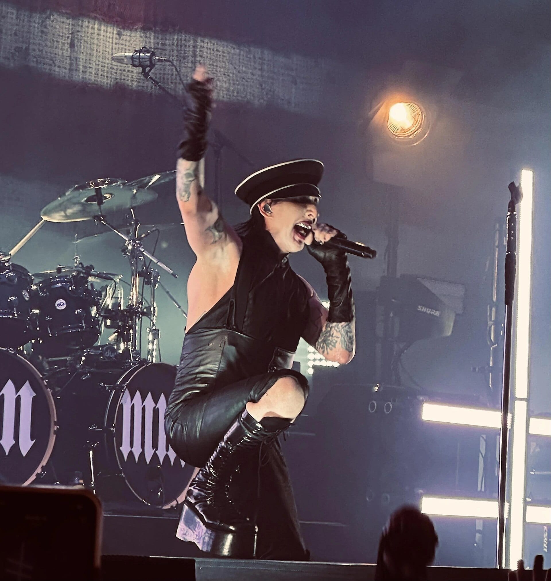 Marilyn Manson leaps mid-performance on stage, wearing a black peaked cap and leather outfit and gripping a microphone, with his band's drum kit visible behind him.