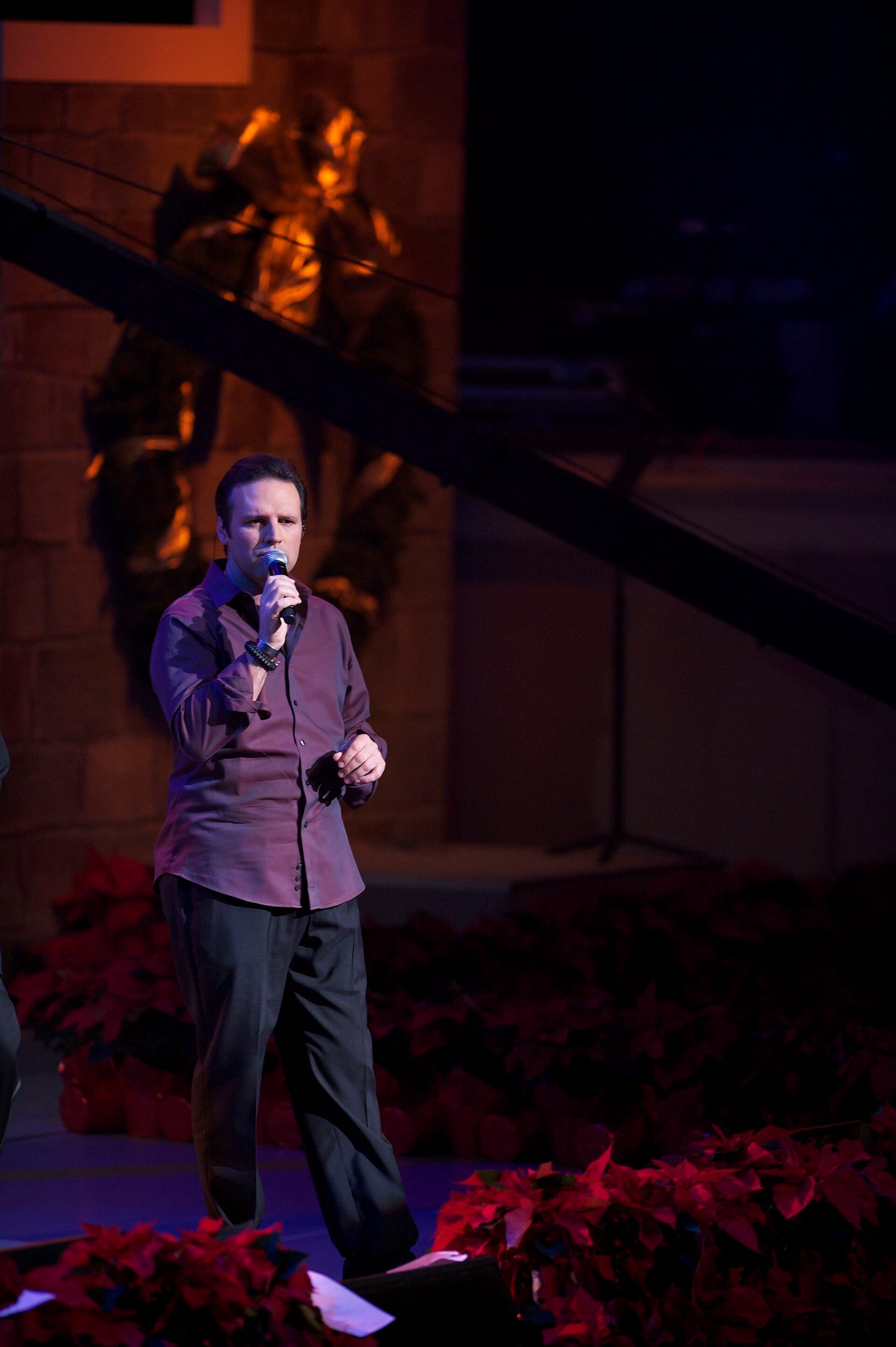 Mark Wills sings into a handheld microphone on a dimly lit outdoor stage, wearing a maroon button-up shirt, with red poinsettias and a lit gold garland behind him.
