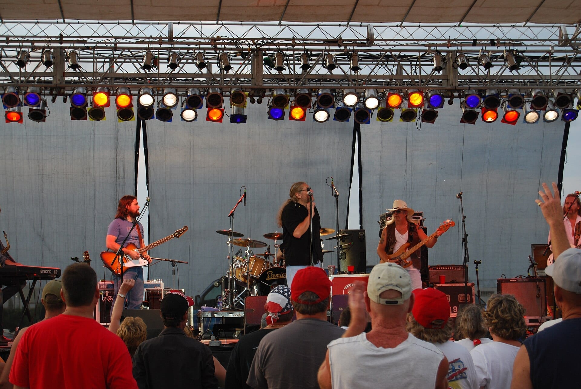 The Marshall Tucker Band play an outdoor stage at dusk under a rack of coloured par cans, a singer at the centre mic with a guitarist to his left and a bassist in a white cowboy hat to his right, seen over the heads of the crowd.