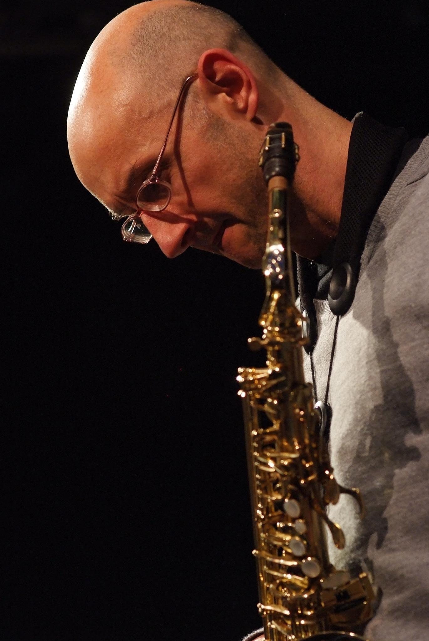 Close-up of jazz saxophonist Martin Speake, bald and wearing glasses, playing a soprano saxophone with his head bowed toward the instrument.