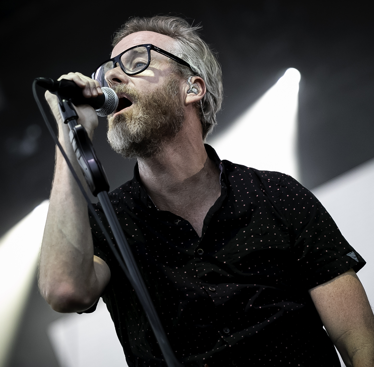 Matt Berninger sings into a handheld microphone with his head tipped back, in dark-framed glasses and a black patterned shirt, stage lights behind him.