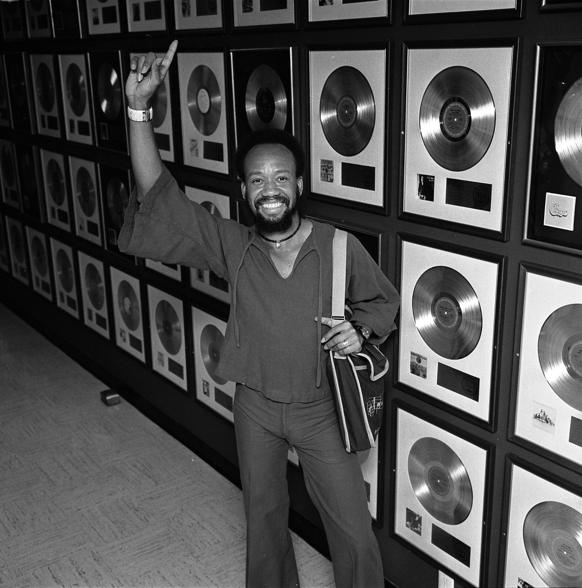A bearded man in a loose shirt grins and points upward while standing in front of a wall covered in framed gold and platinum records, in black and white