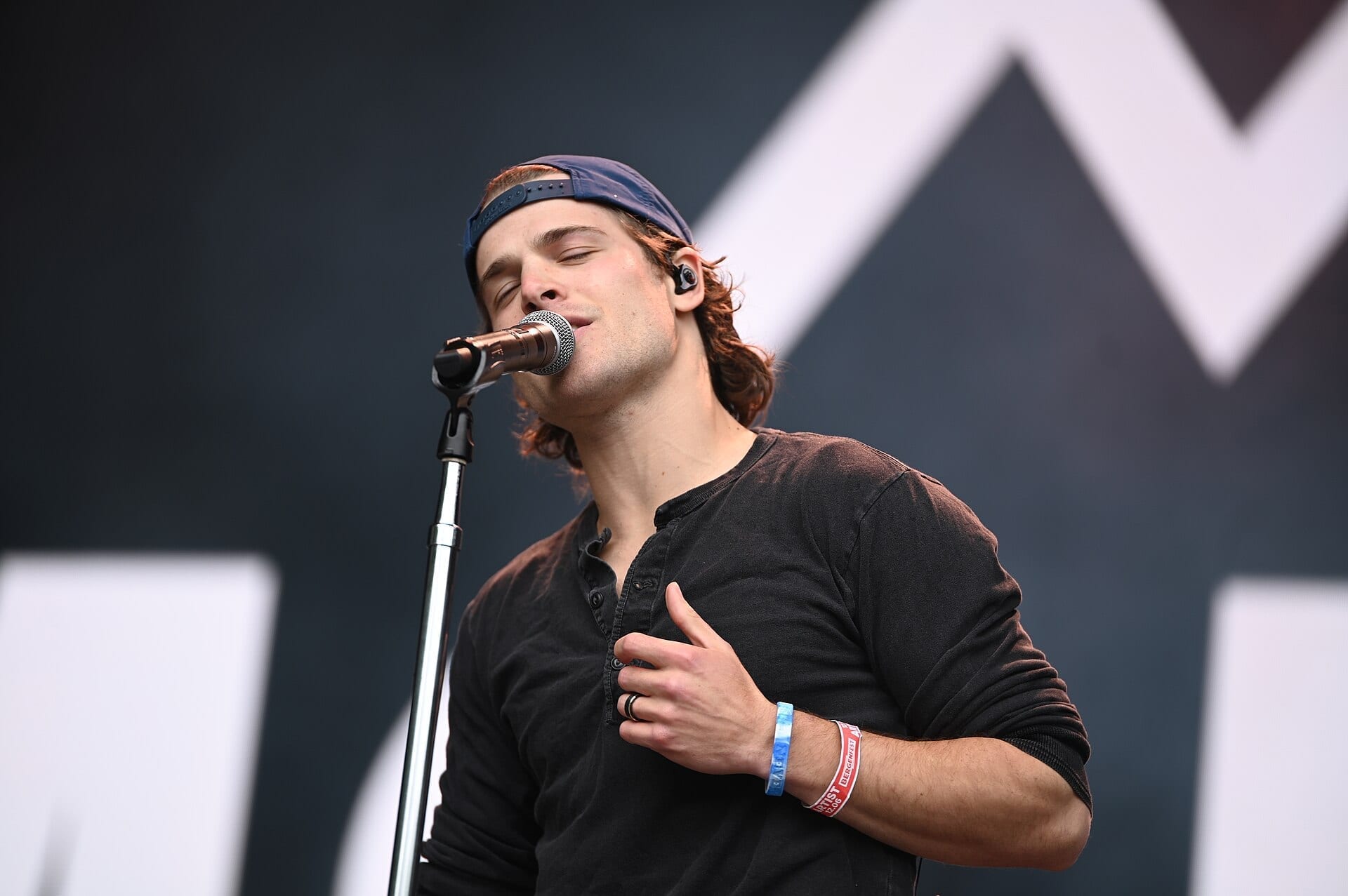 Max McNown sings into a microphone on stage with his eyes closed, wearing a backwards cap and a black long-sleeve shirt.