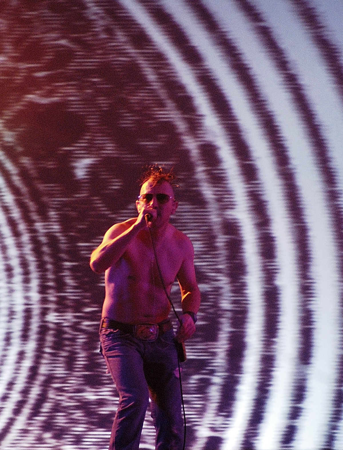 A shirtless singer with a short mohawk and dark sunglasses sings into a handheld microphone in front of a large striped video projection.