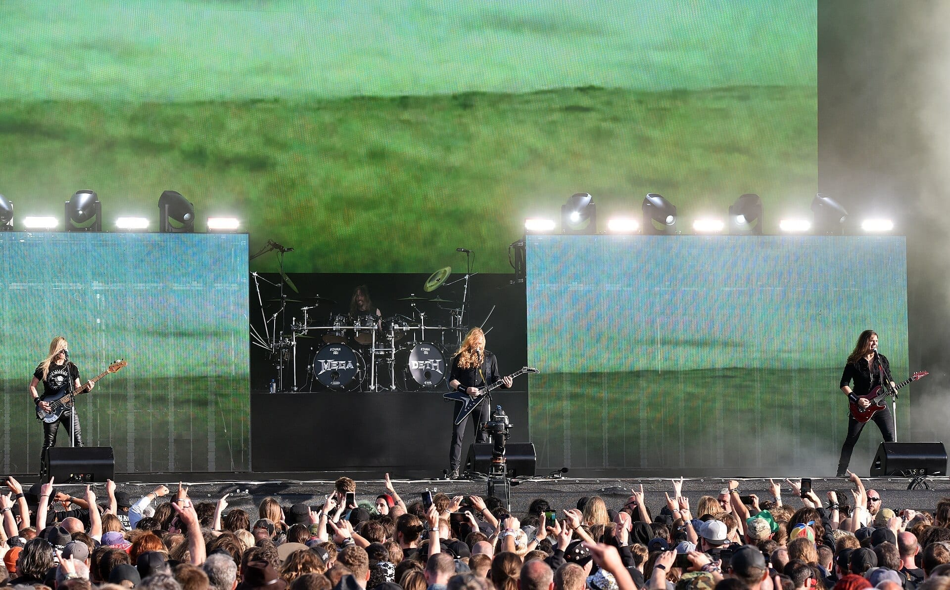 Megadeth performs on a wide outdoor festival stage, guitarists and a bassist flanking a drum kit that reads MEGADETH, in front of a large crowd.