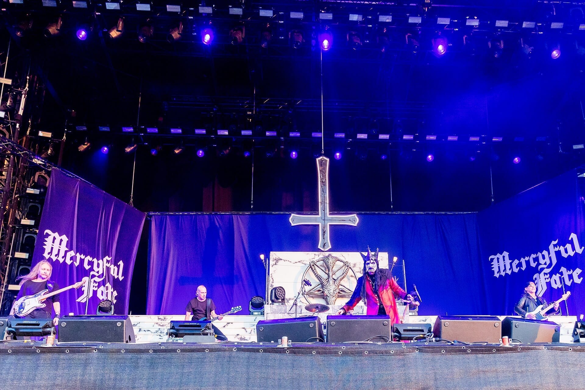 Mercyful Fate performs on a large festival stage beneath an inverted cross and pentagram backdrop, with singer King Diamond in skull face paint at center and the band's name lit up on banners on either side.
