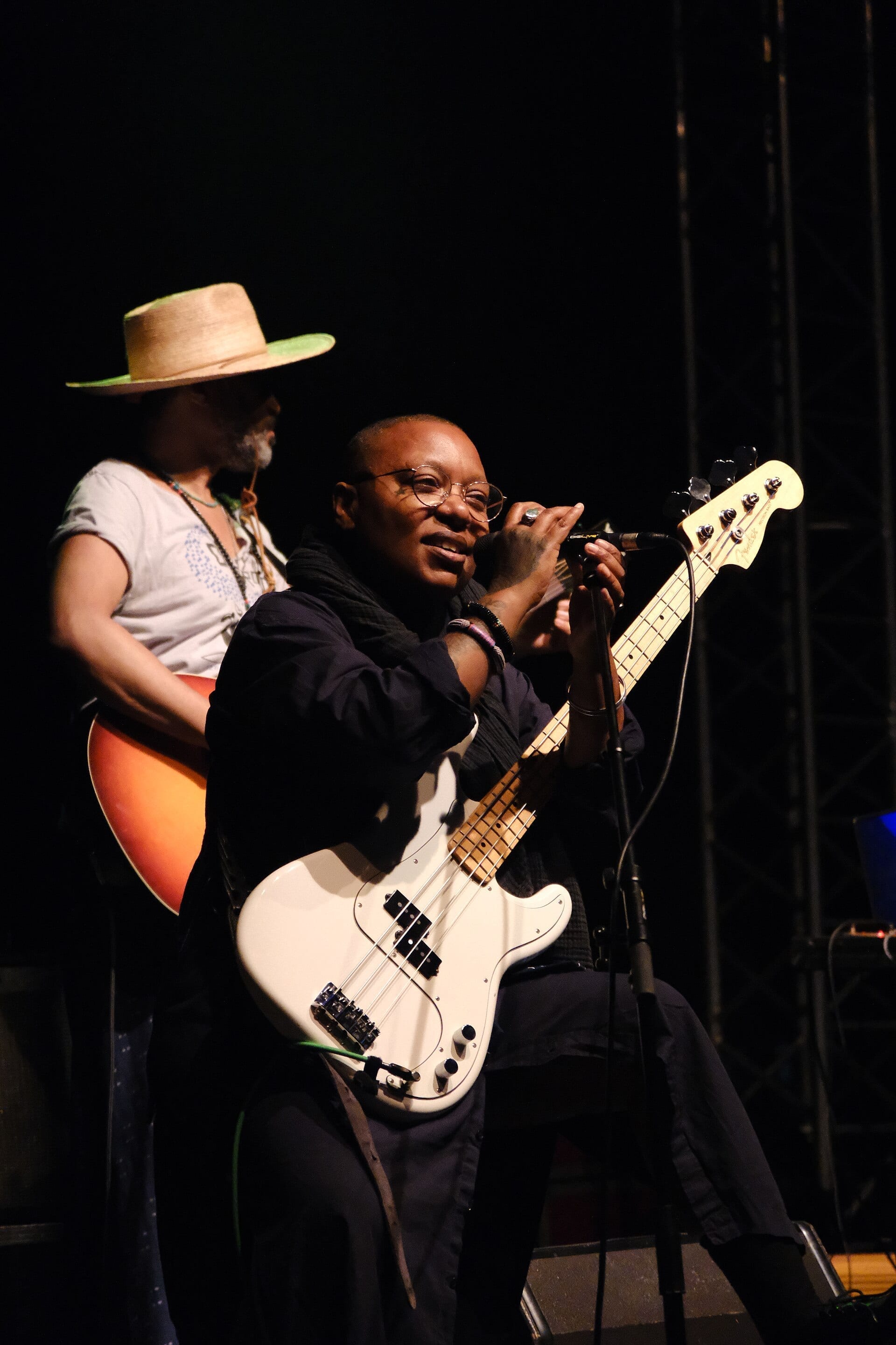 Meshell Ndegeocello, bald and wearing round glasses, plays a white bass guitar and sings into a microphone on stage, with a guitarist in a straw hat behind her.