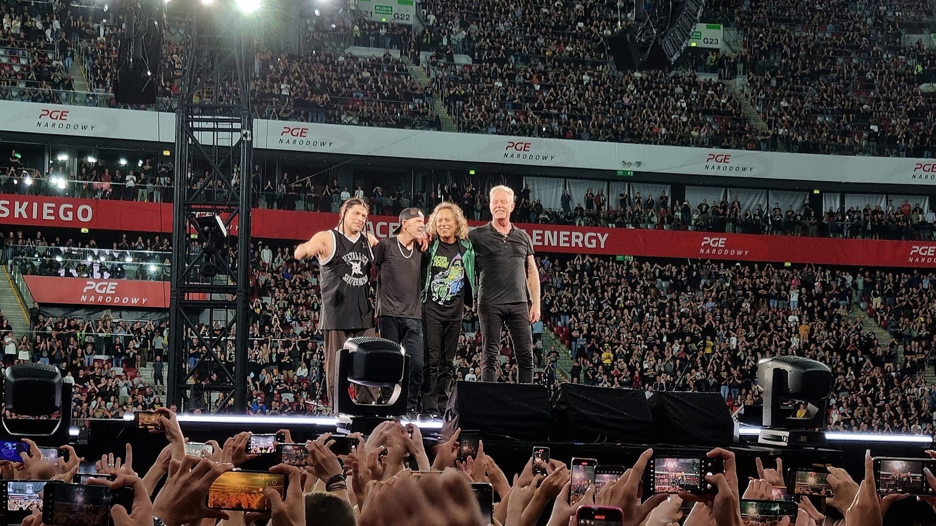 The four members of Metallica stand together on stage waving to a packed stadium crowd, many fans holding up phones to record.