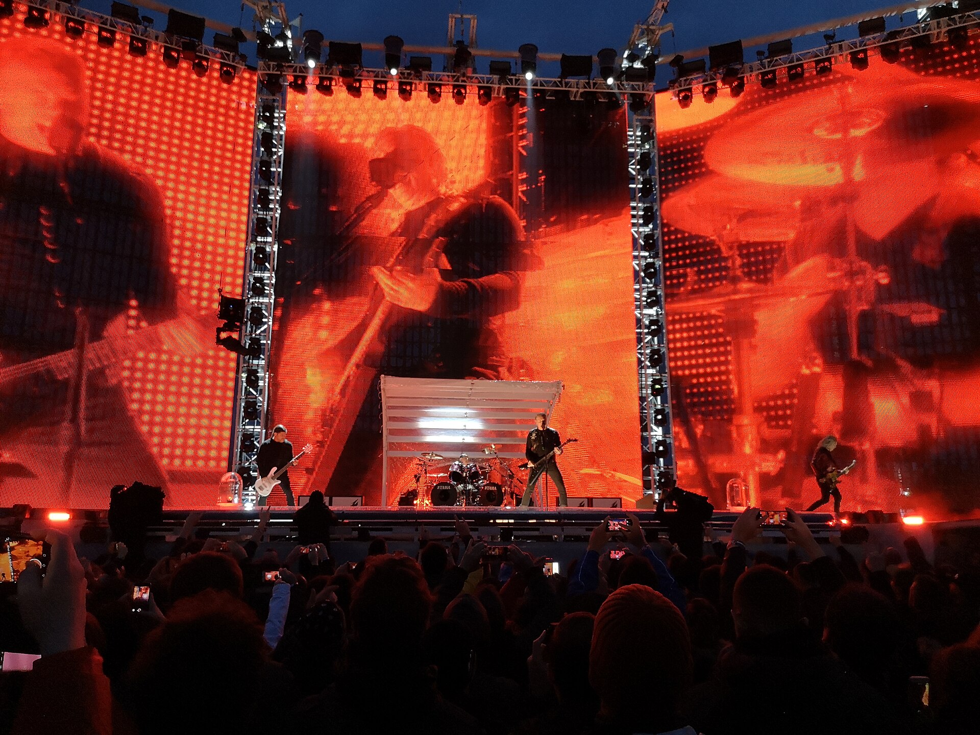 Four musicians spread across a wide stadium stage in front of towering red video screens, a crowd holding up phones in the foreground