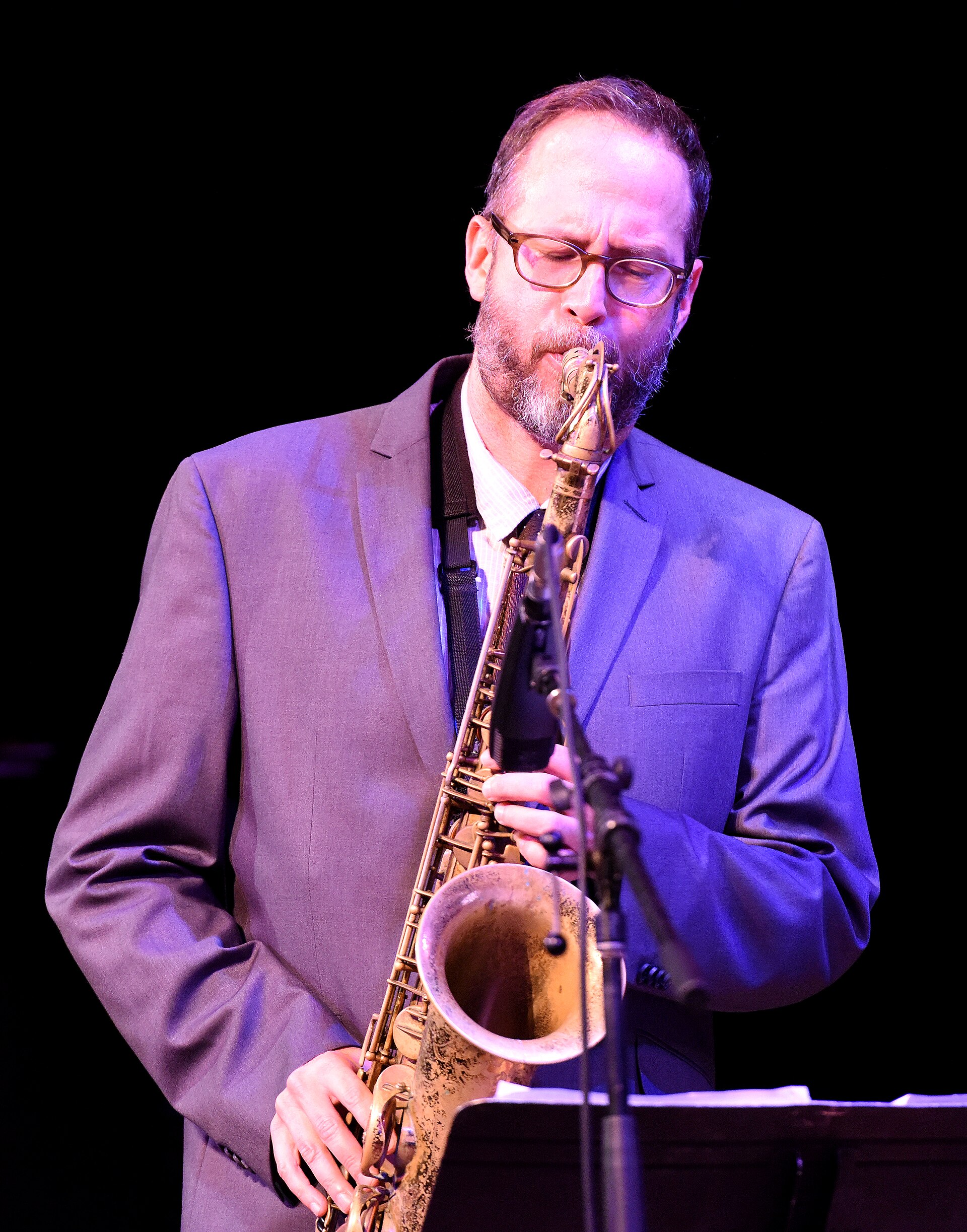 Saxophonist Michael Blake plays a tenor saxophone in a dark suit, lit from the side on a concert stage.