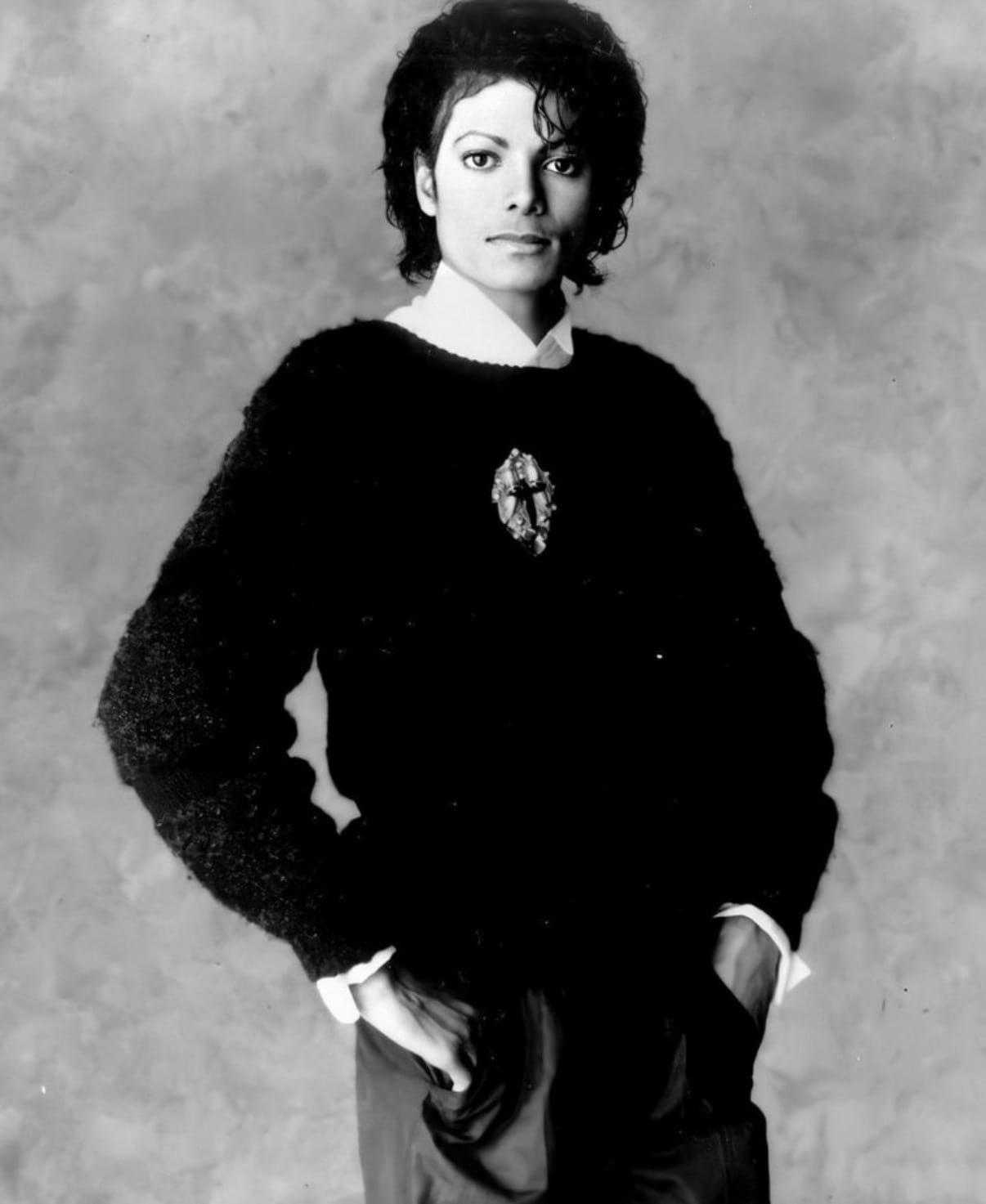 A black-and-white studio portrait of Michael Jackson wearing a dark sweater with a jeweled brooch, hands in his pockets, looking at the camera.