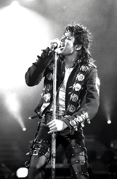 Michael Jackson sings into a microphone stand on stage, wearing his signature military-style studded jacket, in this black-and-white photo.