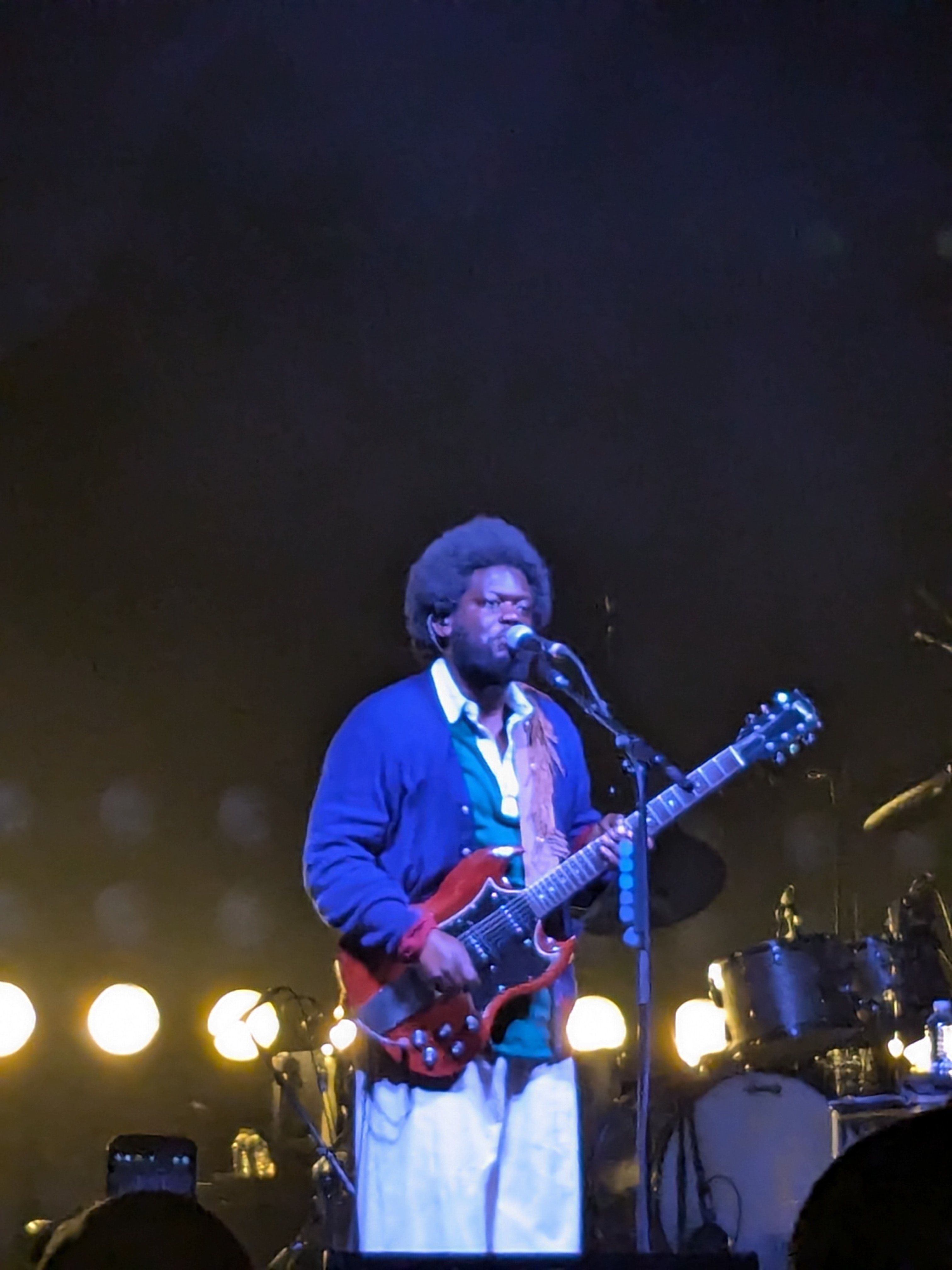 Michael Kiwanuka sings into a microphone while playing a red electric guitar, wearing a blue cardigan over a shirt, lit blue on a dark stage.