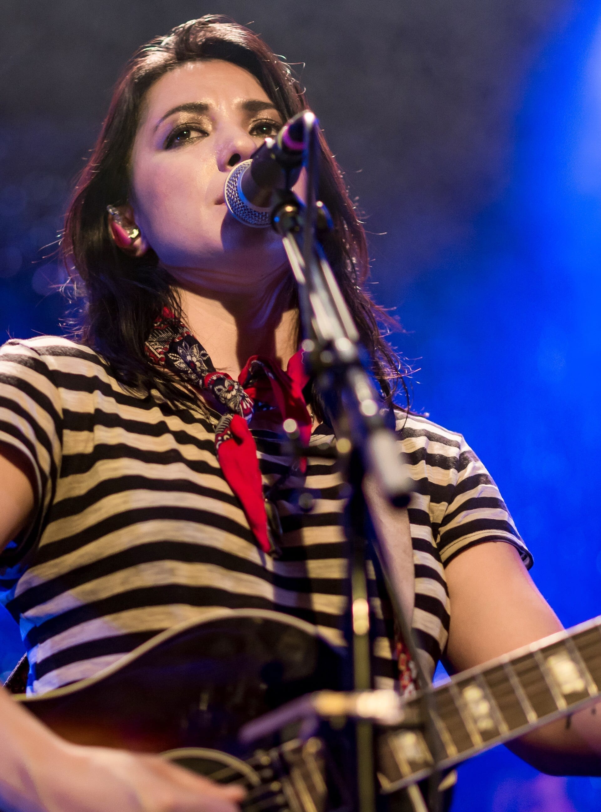Michelle Branch, wearing a striped top and a red bandana, sings into a microphone while playing guitar under blue stage light.