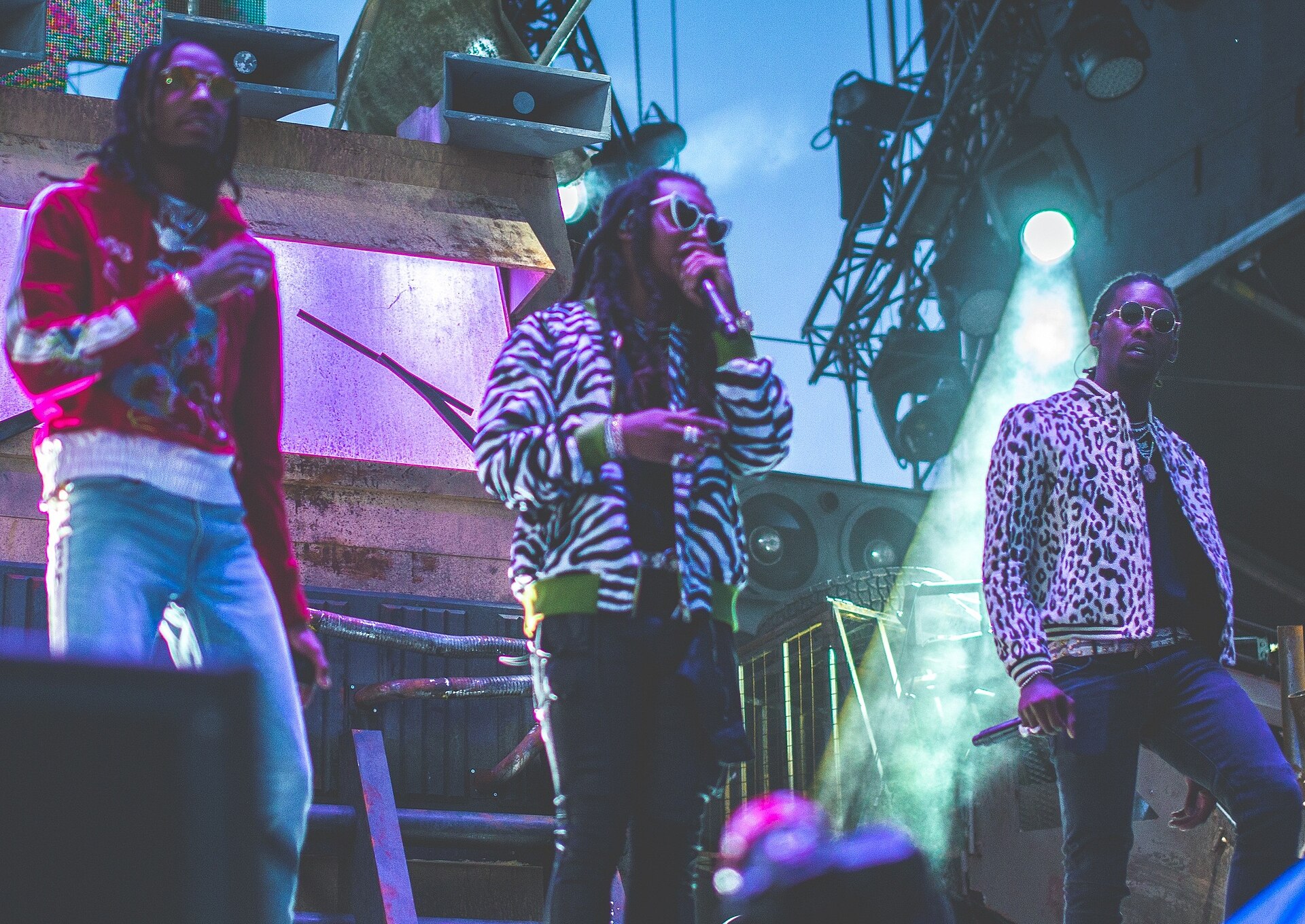 Three rappers spread across an outdoor festival stage in sunglasses and patterned jackets, one at the microphone, stage rigging and a spotlight behind them