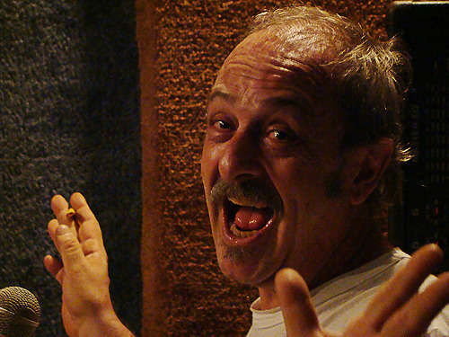 Miguel Zavaleta sings with his mouth wide open and both hands raised, close up in a warmly lit room.