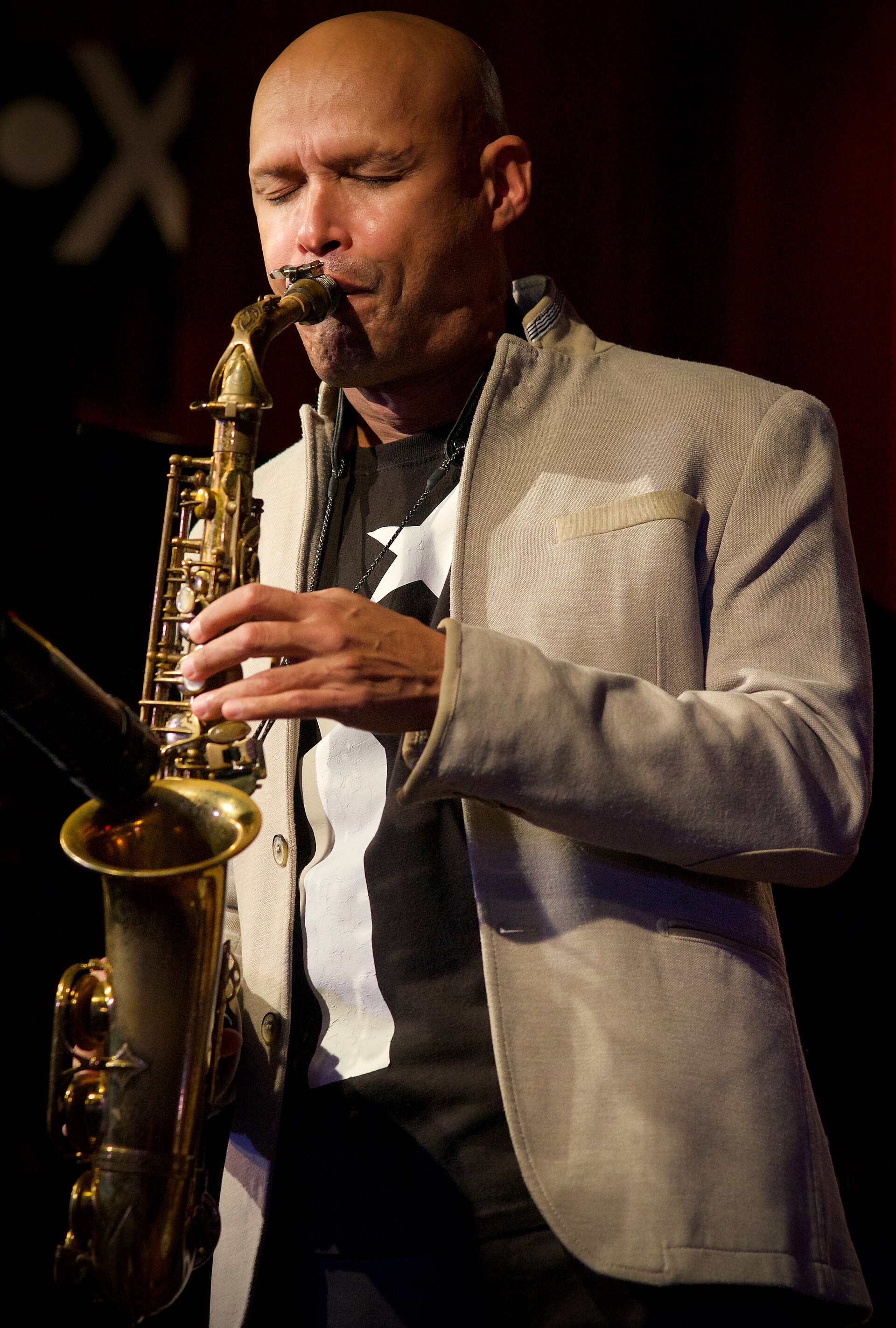 Miguel Zenon plays alto saxophone with his eyes closed, wearing a light jacket over a printed shirt.