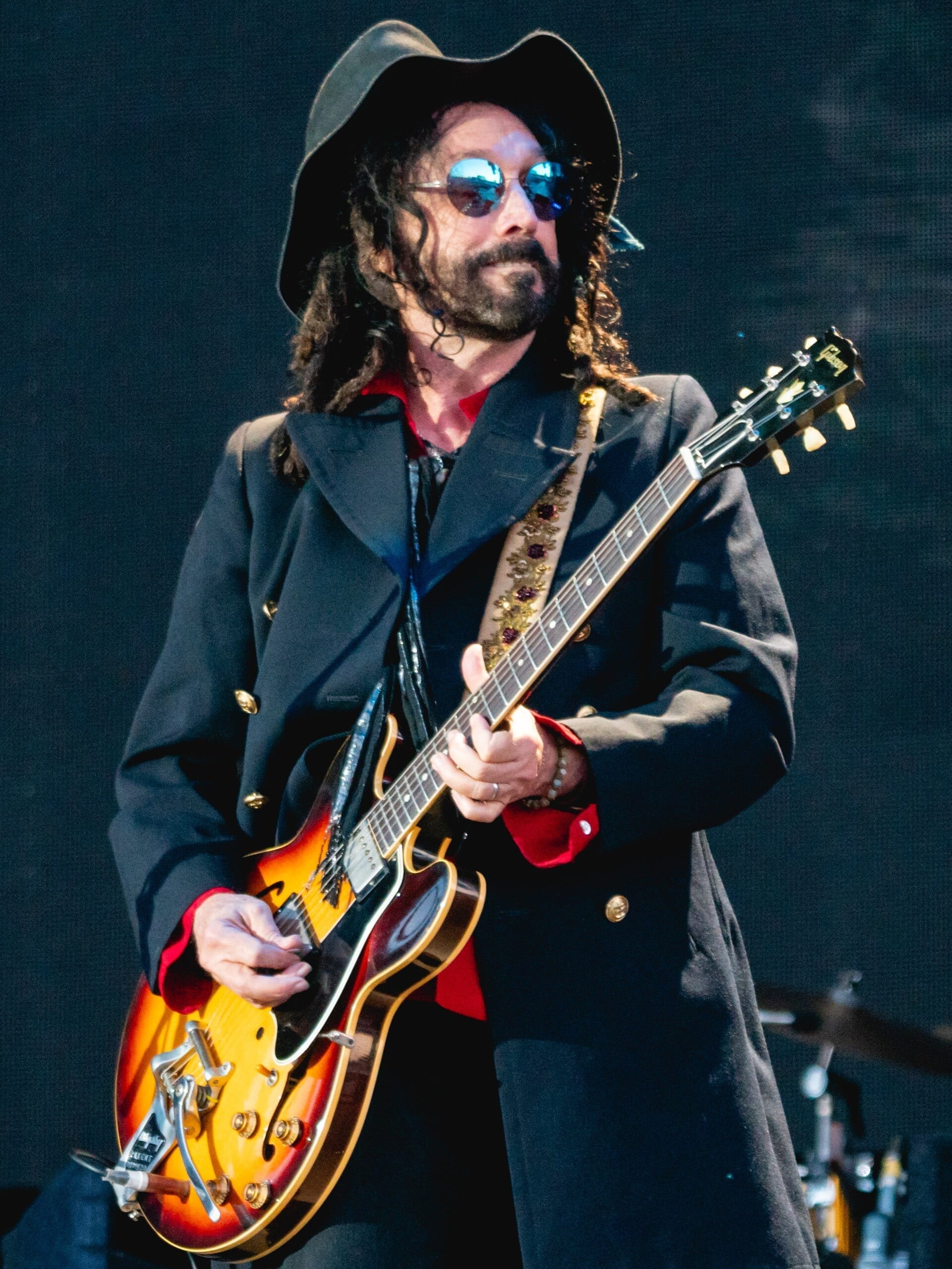 Mike Campbell, wearing a wide-brimmed hat and mirrored sunglasses, plays a sunburst electric guitar on stage.
