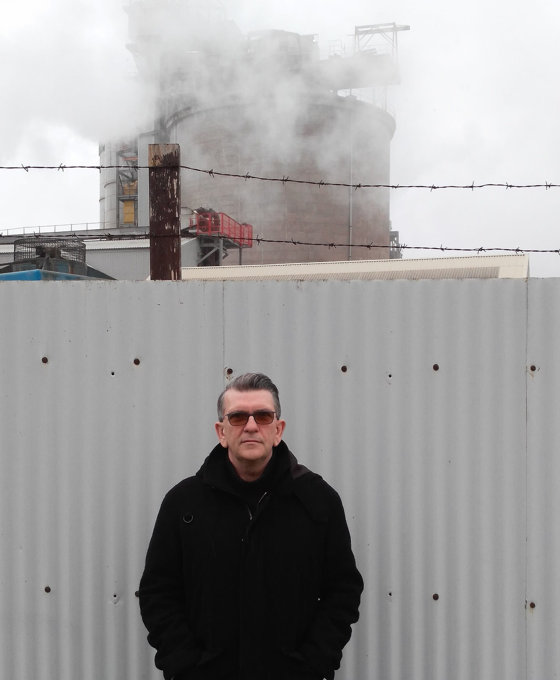 Mike Joyce stands in sunglasses and a black jacket in front of a corrugated metal fence, with an industrial cooling tower and rising steam behind him.
