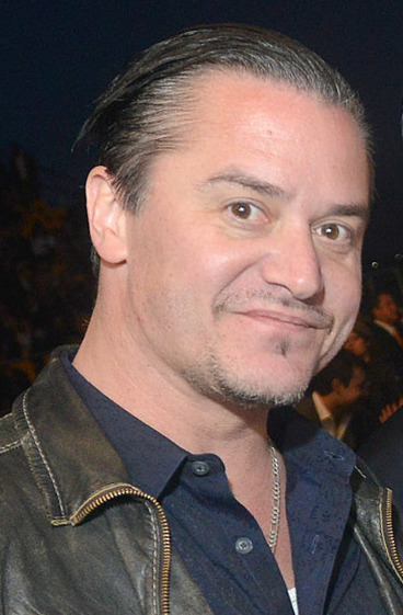 Mike Patton half-smiles at the camera, hair slicked back, in a leather jacket over a dark blue shirt and a thin chain.