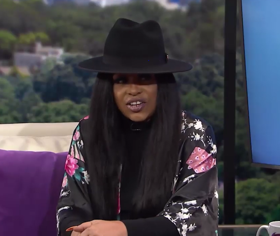 Miki Howard sits on a talk show set in a black fedora and a floral bomber jacket, long straight hair over one shoulder, mid-sentence.