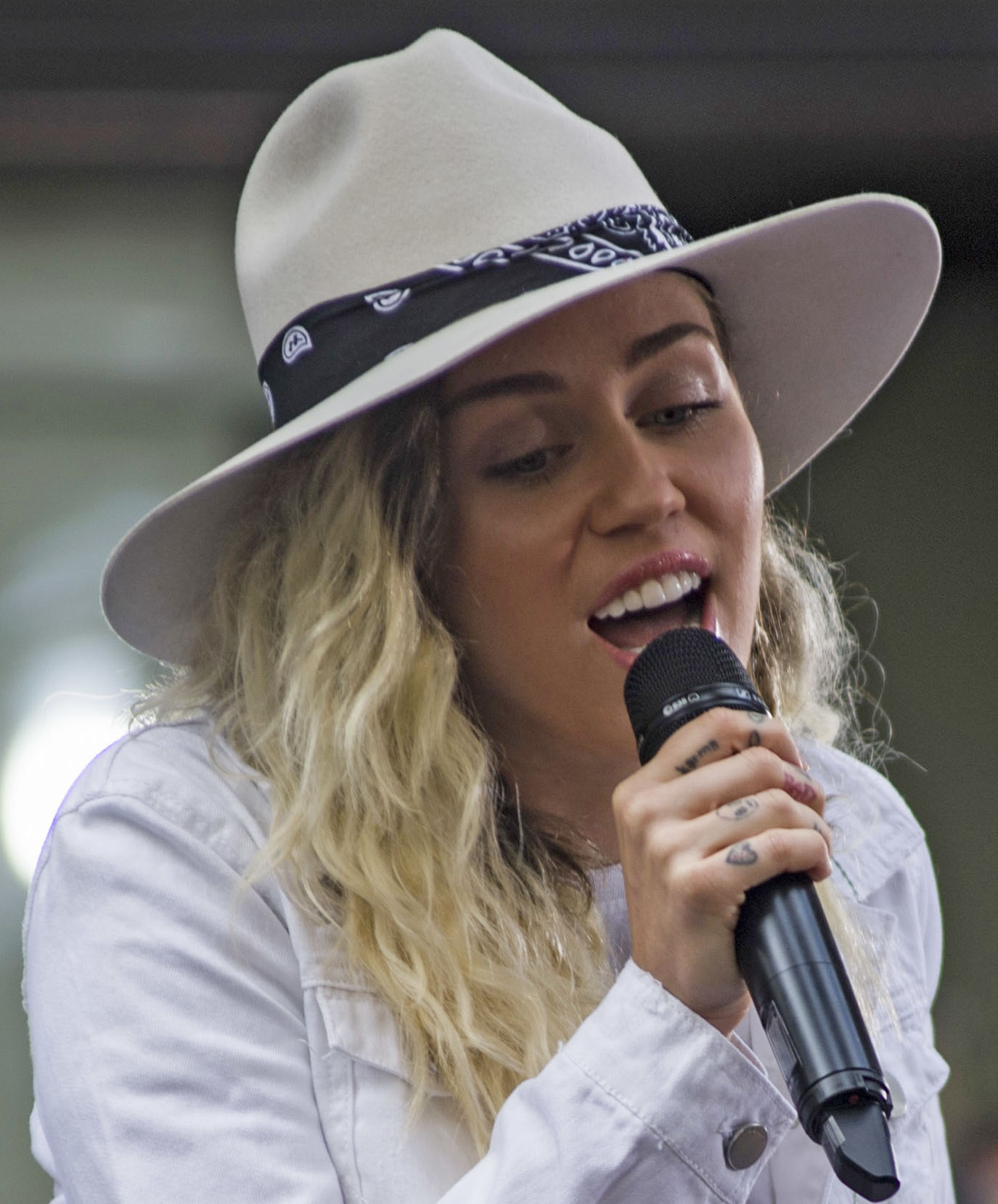 Miley Cyrus sings into a black handheld microphone in a wide cream hat with a bandana band and a white denim jacket.