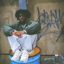 Millkzy sits on an overturned blue barrel in front of a graffiti-covered wall, wearing a green jacket, white pants and white sneakers, arms resting on his knees.