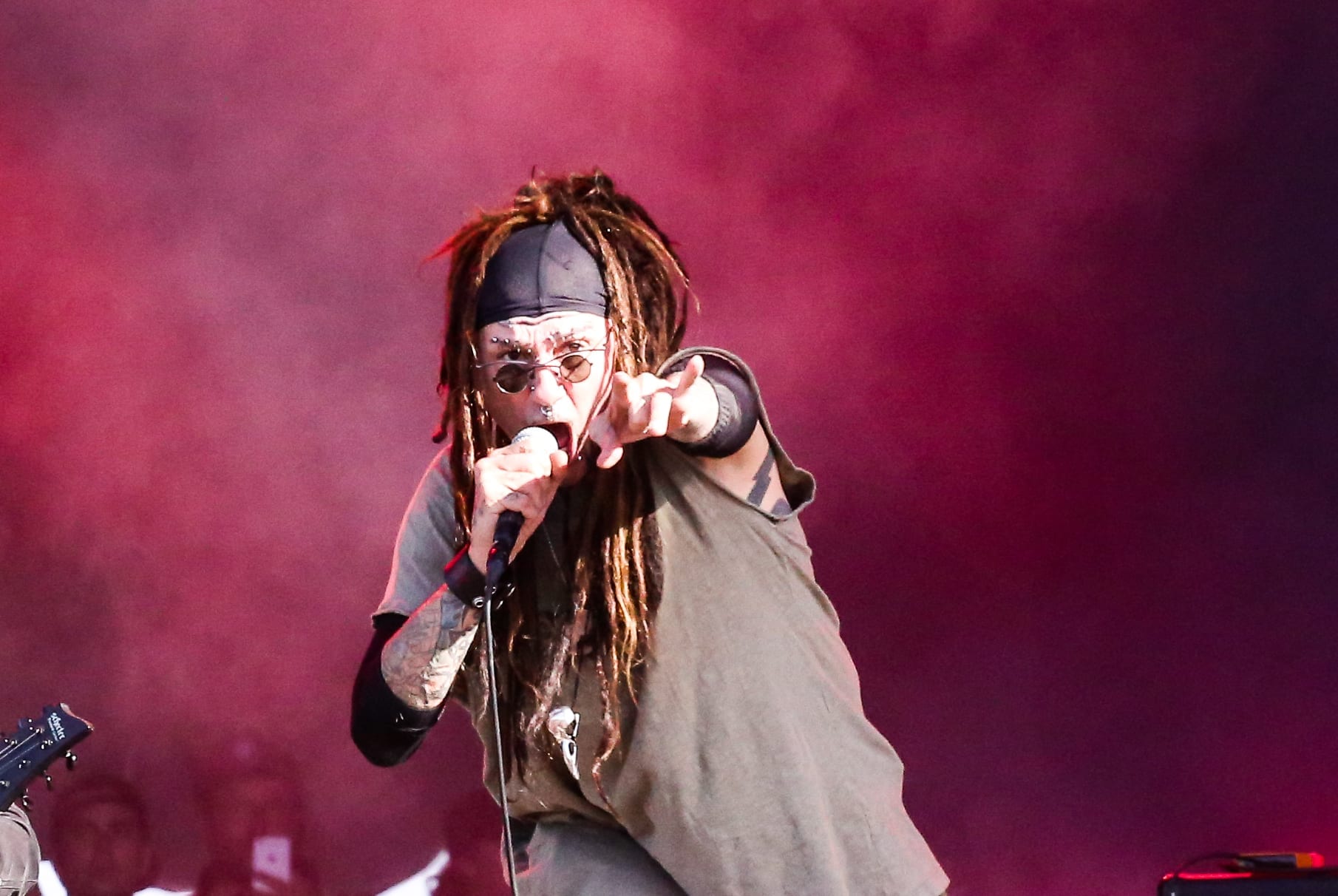 Al Jourgensen shouts into a microphone and points at the crowd, dreadlocks under a black bandana and small round sunglasses, against red haze.