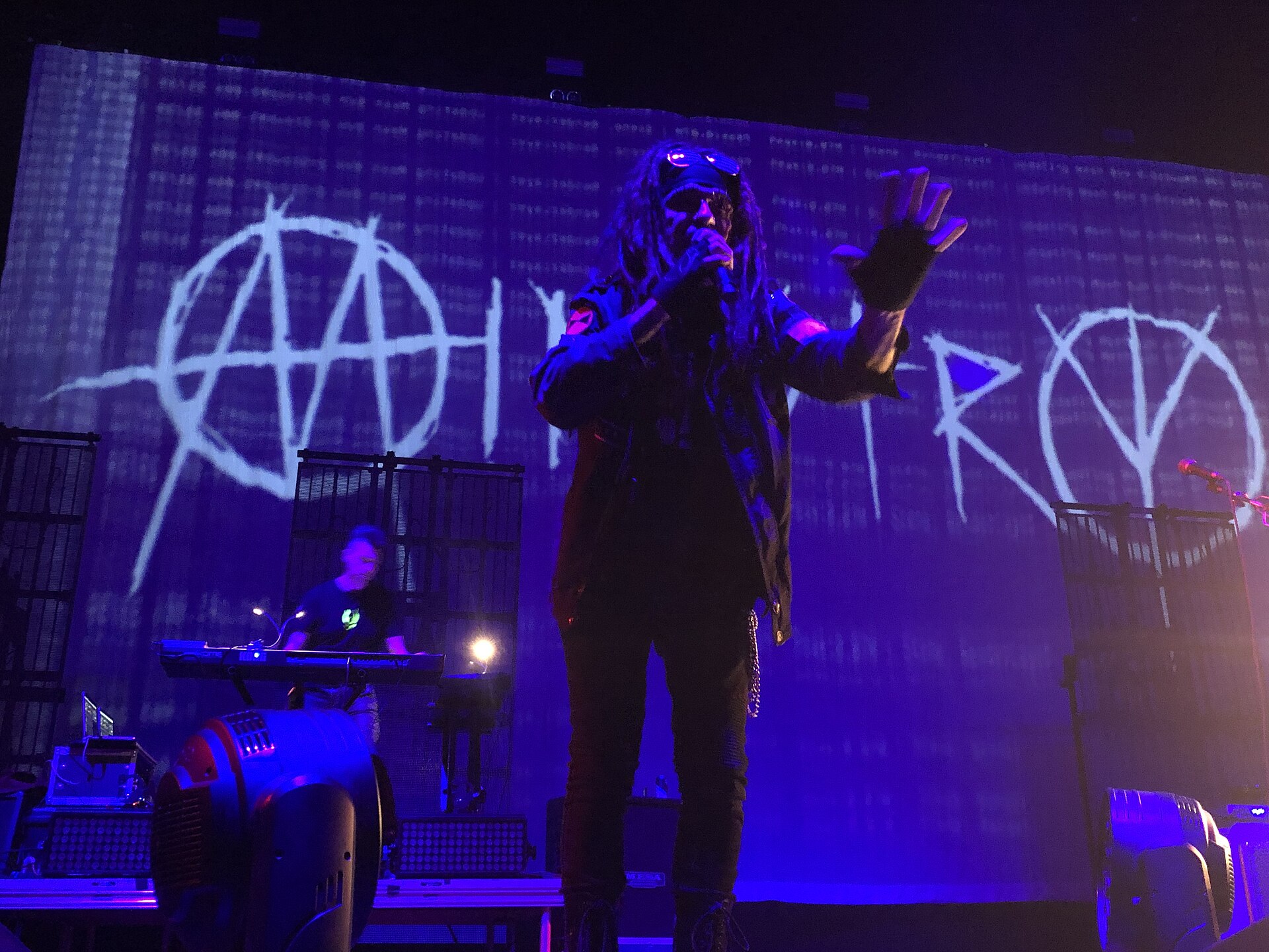 A singer in dreadlocks and goggles holds a microphone with one hand raised, lit blue in front of a backdrop painted with the band name Ministry, a keyboard player behind him