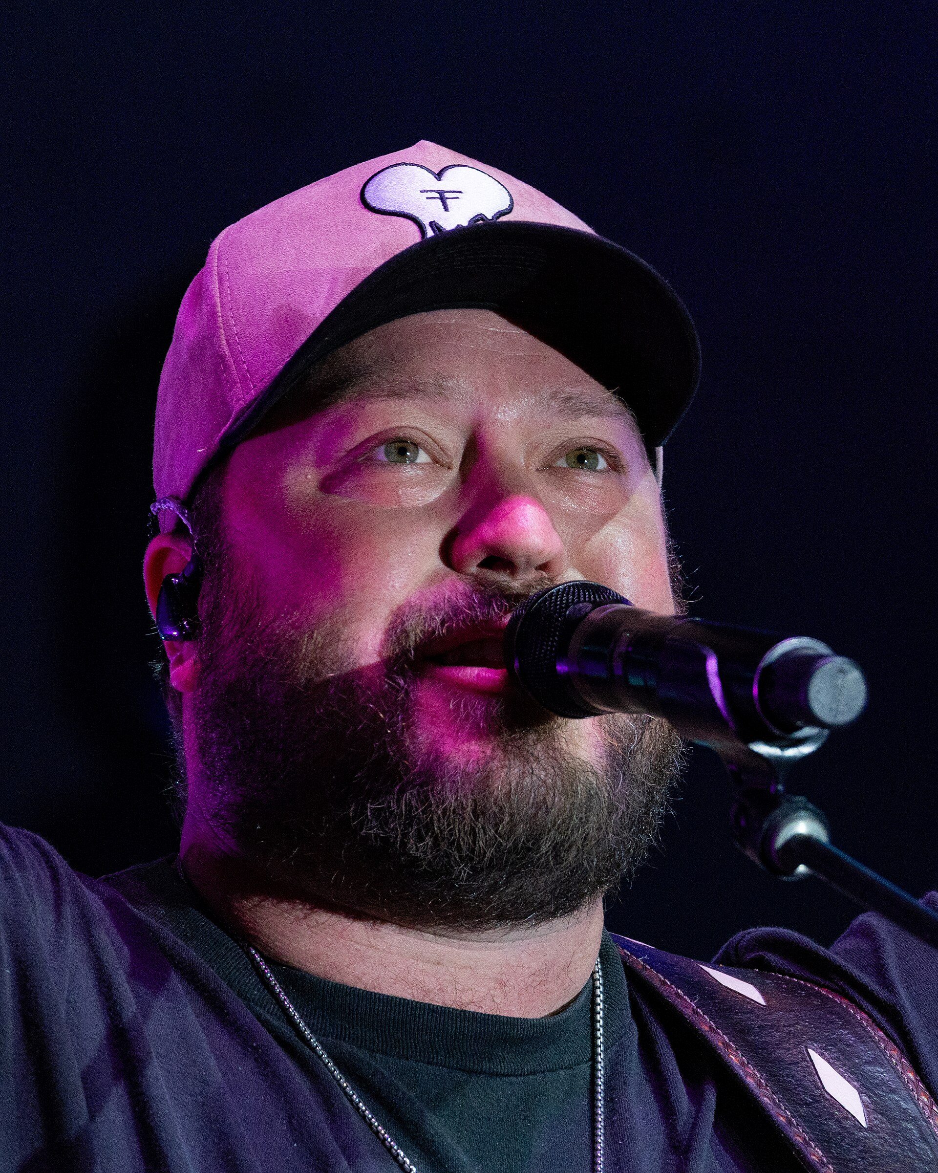 Mitchell Tenpenny sings into a microphone in close-up under pink stage light, full beard, an in-ear monitor in place and a pink cap turned forward.