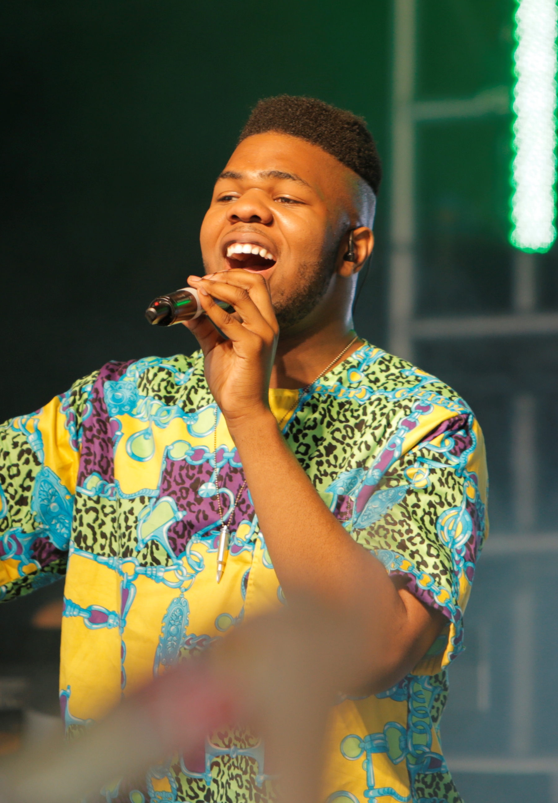 MNEK sings with his eyes closed, holding a microphone to his mouth in a bright yellow, green and blue patterned shirt, green stage lights behind him.