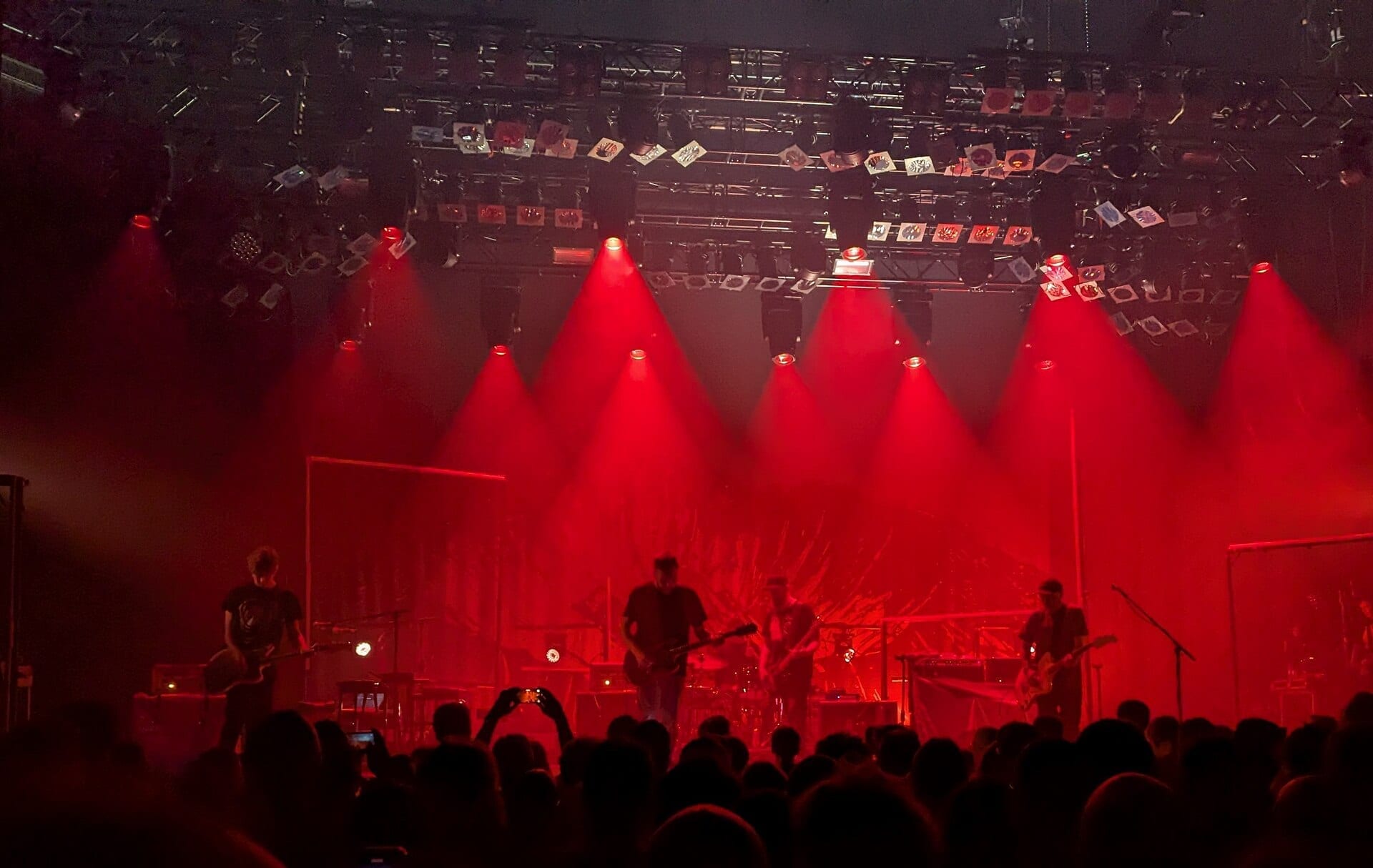 Mogwai's four members play guitars and keyboards in silhouette on a red-lit stage, framed by hanging light rigs, with a crowd watching in the foreground.