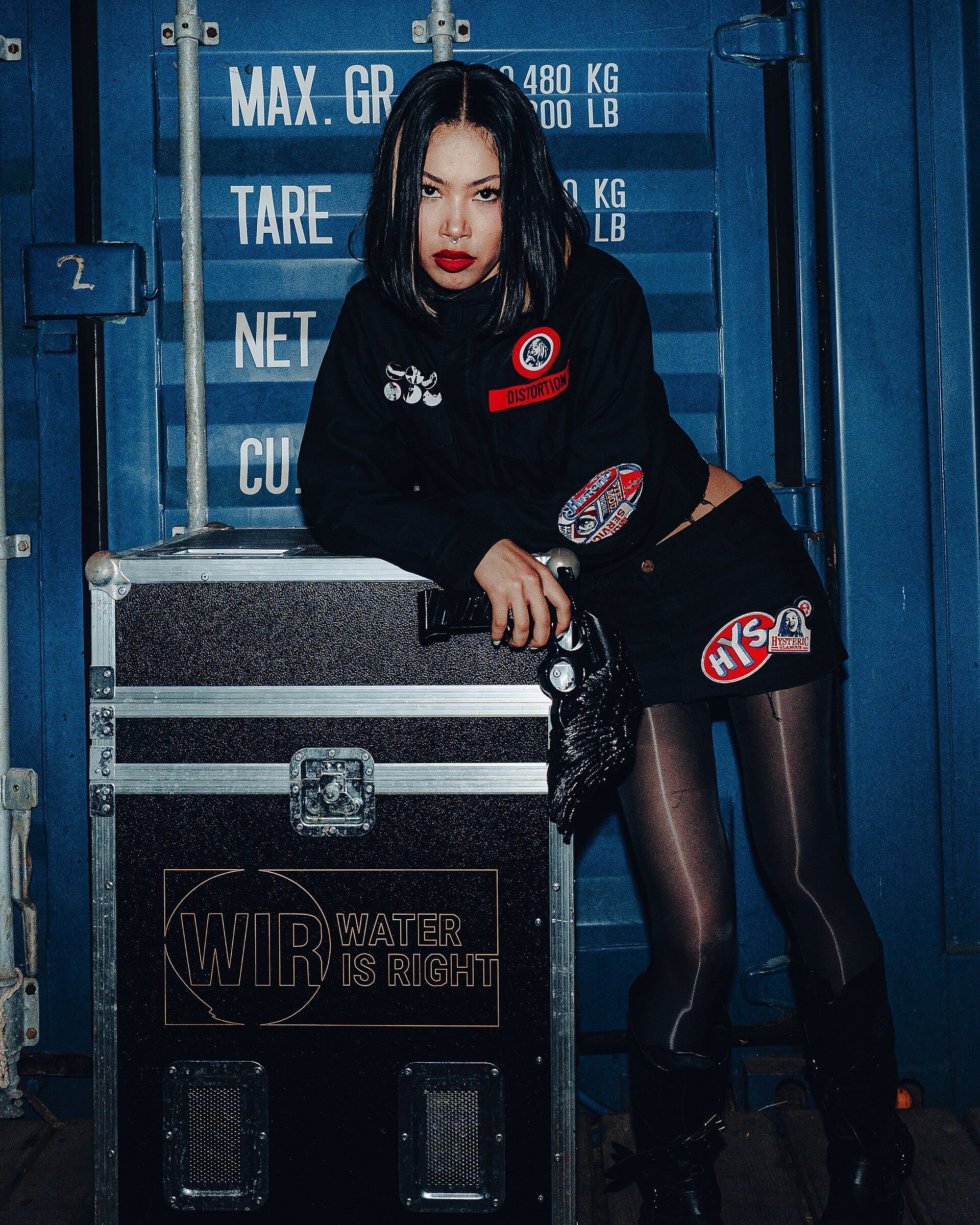 Molly Santana leans on a road case backstage, wearing a black jacket and skirt covered in patches, with dark hair and red lipstick.