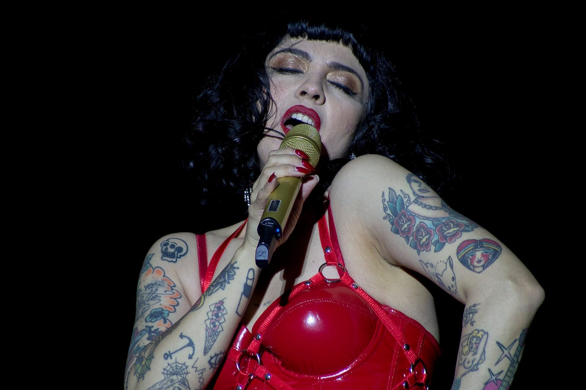 Mon Laferte sings into a gold microphone with eyes closed, wearing a red latex top that shows her many tattoos.