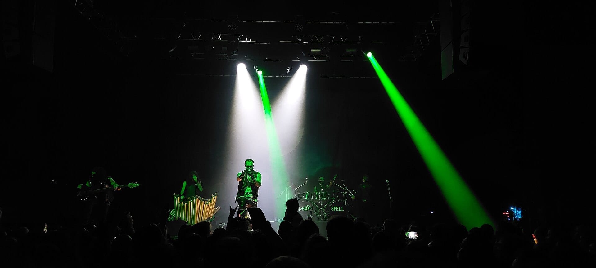 The singer of Moonspell stands at center stage under crossing green and white spotlights, backed by bandmates and a lit MOONSPELL sign, as a crowd raises their phones below.