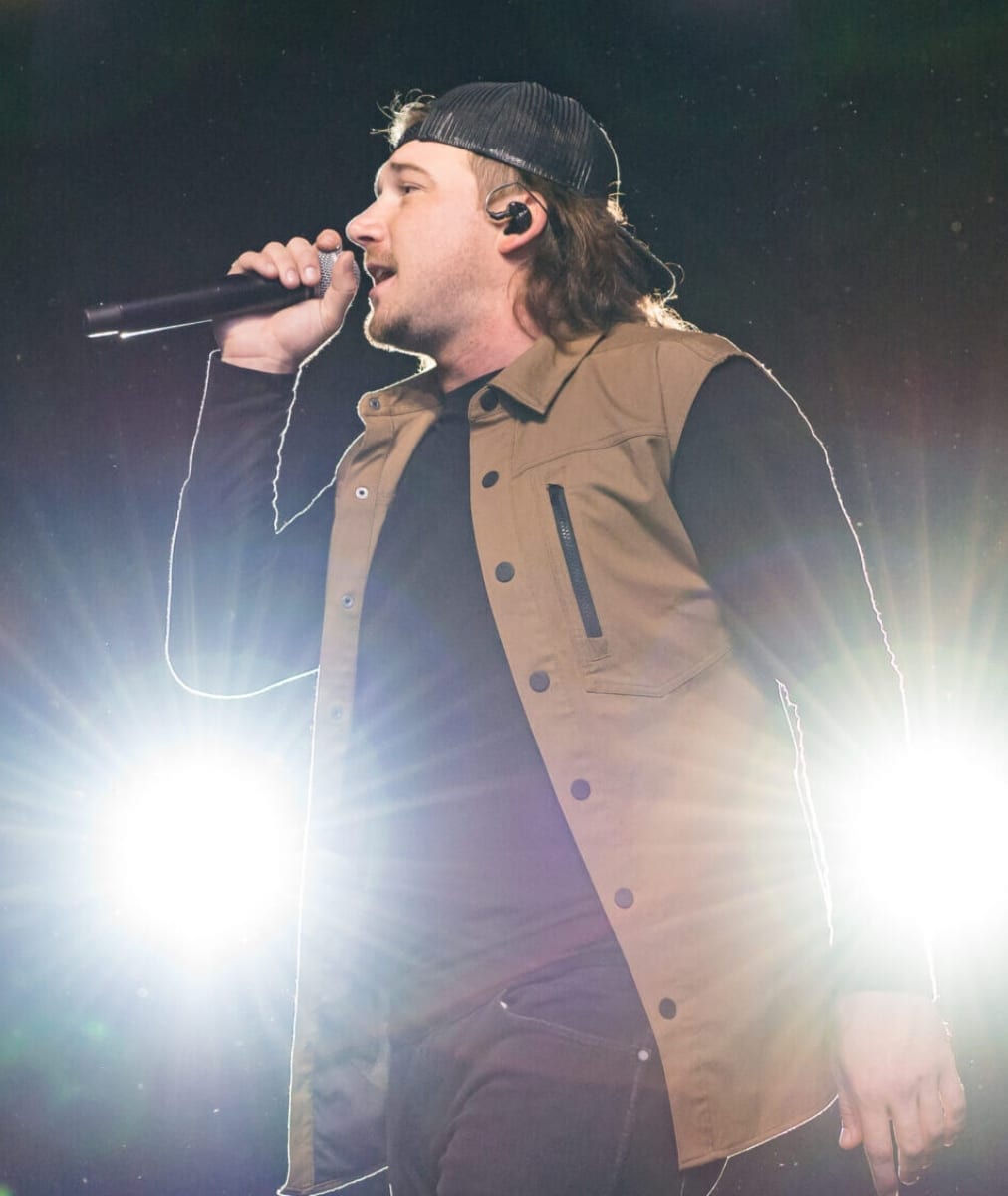 Morgan Wallen sings into a handheld microphone on a brightly backlit stage, wearing a backward black beanie and a tan vest over a black shirt.