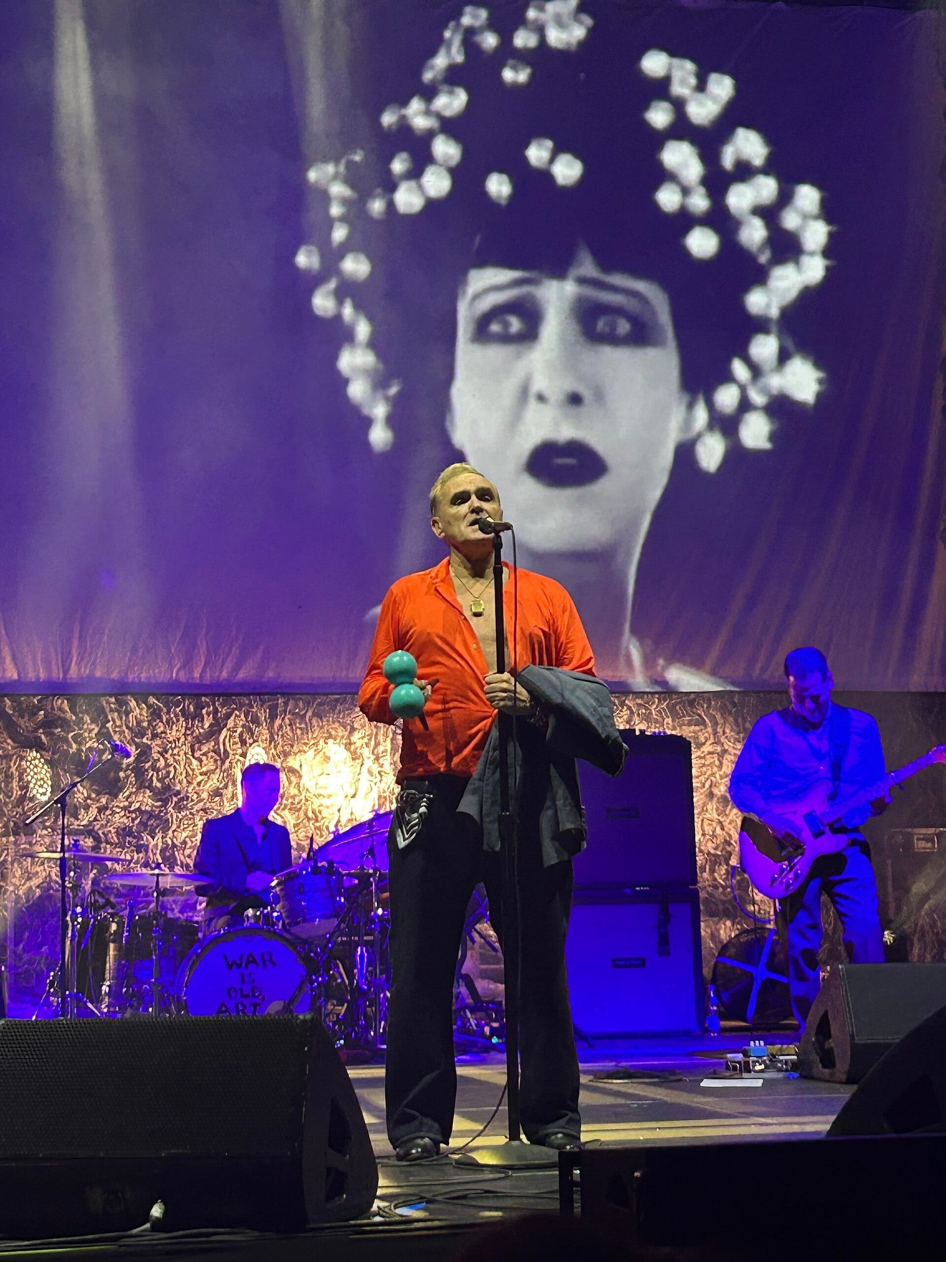 Morrissey sings into a microphone in an open orange shirt, holding maracas, in front of a giant black and white backdrop of a woman with an elaborate flowered headpiece, band members behind him.